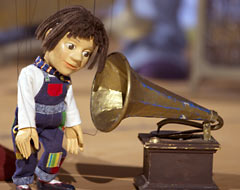 Marionette Paula hält ihr Ohr an ein Grammofon.