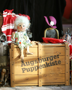 Die Koboldmarionette Freudibold sitzt auf einer Kiste, die den Schriftzug Augsburger Puppenkiste trägt.
